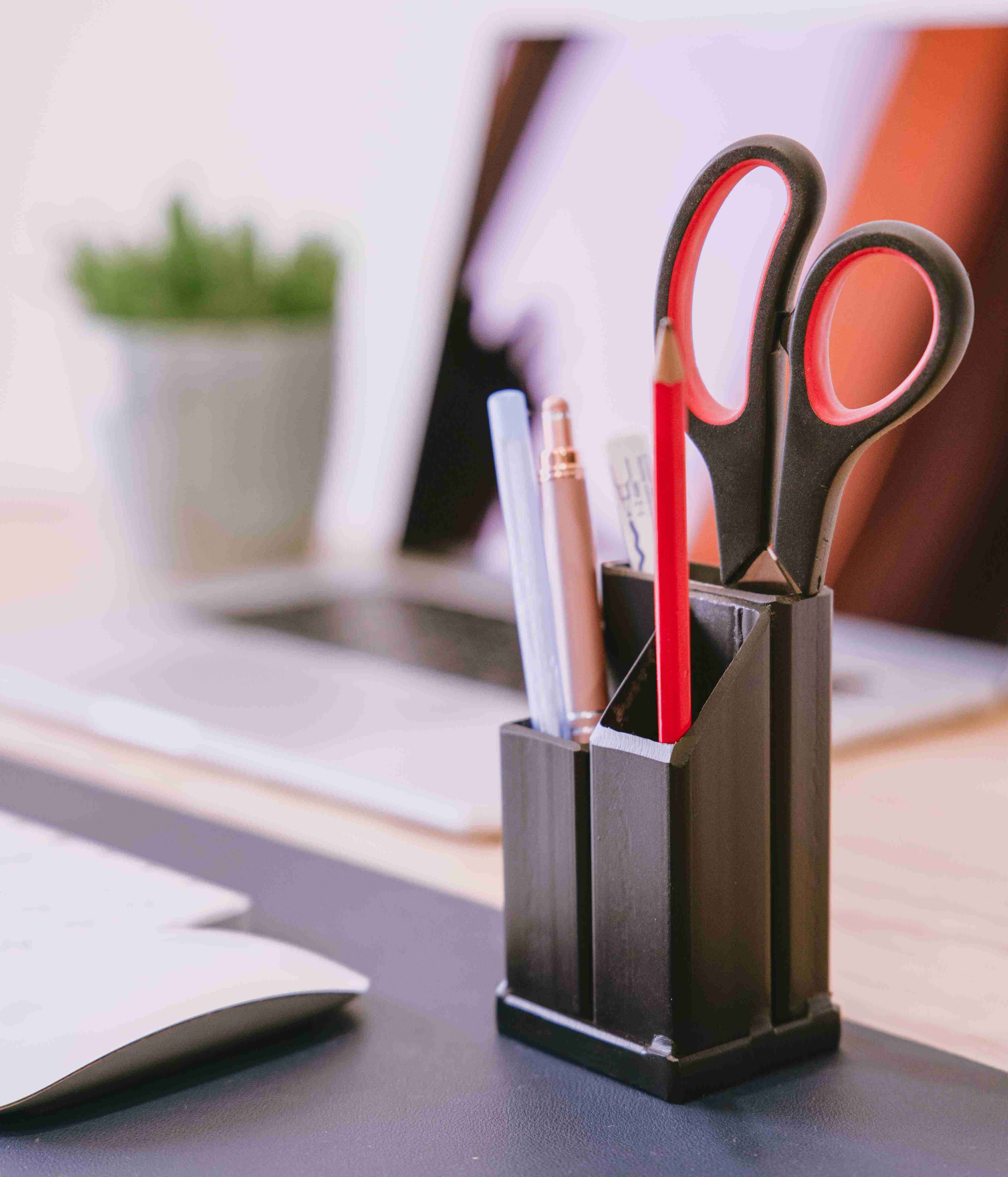 Matt black steel pen holder with pens pencils and scissors inside of it on a desk with a laptop and a plant in the background.