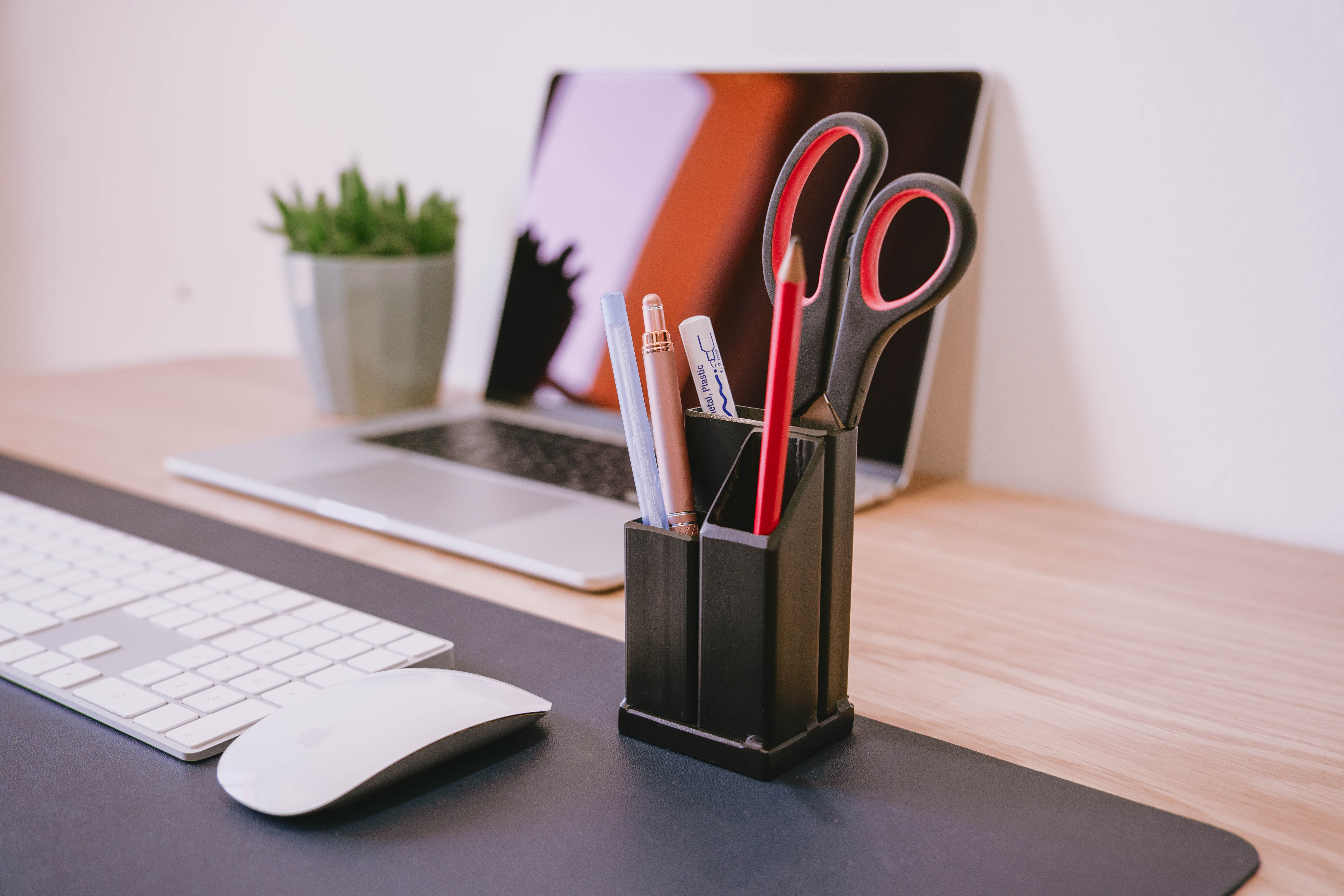 Matt black steel pen holder with pens pencils and scissors inside of it on a desk with a laptop and a plant in the background.